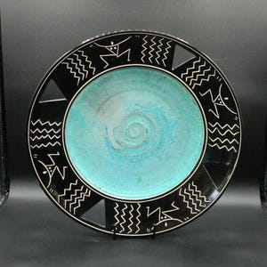 Puede incluir: Un plato grande y redondo de cerámica con un esmalte turquesa y un diseño geométrico en blanco y negro. El plato tiene un patrón en espiral en el centro y recortes alrededor del borde.