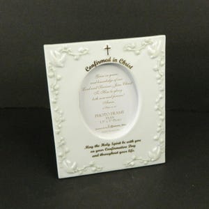 May include: A white ceramic rectangular photo frame with an oval center opening. The frame features embossed dove and vine details, a gold cross at the top, and gold text reading Confirmed in Christ and May the Holy Spirit be with you.