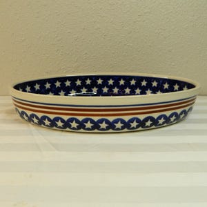 May include: Oval ceramic serving dish with a blue and white star pattern on the inside. The rim features a repeating pattern of blue stars and white circles, with two red stripes. A decorative piece for serving.