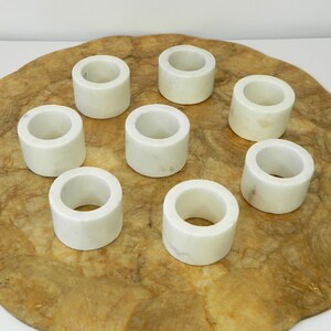 Puede incluir: Ocho servilleteros de m&aacute;rmol blanco. Cada anillo es cil&iacute;ndrico con una abertura circular. Est&aacute;n colocados sobre una superficie ovalada de madera clara, con un veteado natural de color marr&oacute;n.