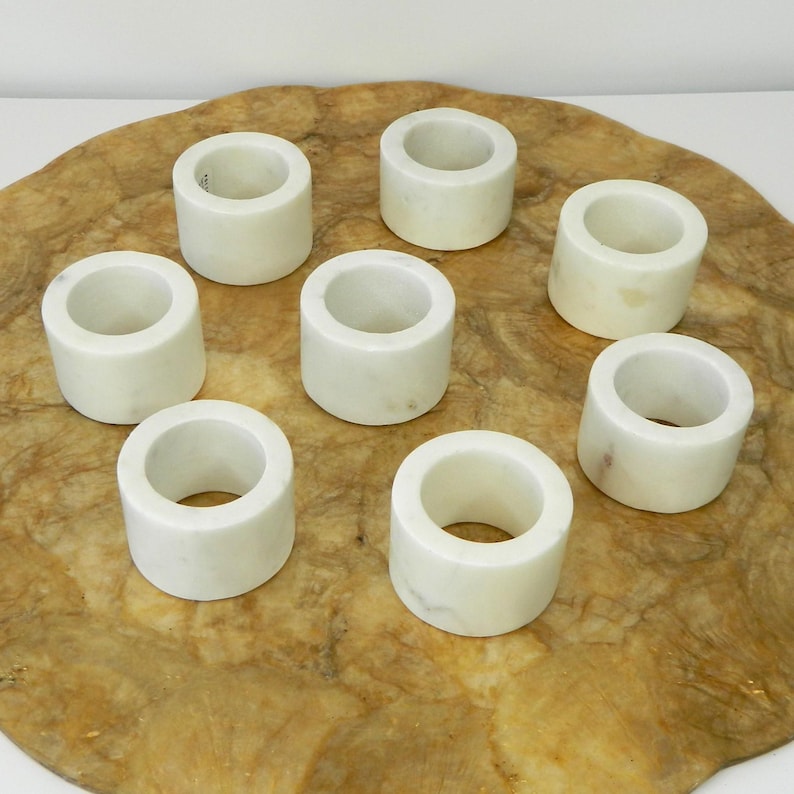 Puede incluir: Ocho servilleteros de m&aacute;rmol blanco. Cada anillo es cil&iacute;ndrico con una abertura circular. Est&aacute;n colocados sobre una superficie ovalada de madera clara, con un veteado natural de color marr&oacute;n.