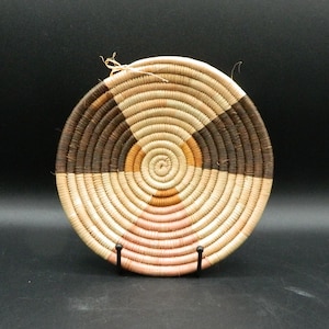 May include: A round woven basket with a brown, beige, and pink geometric pattern. The basket is on a black stand.