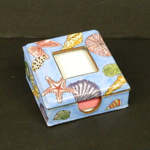May include: A rectangular, light blue trinket box with a hinged lid. The box is decorated with colorful seashells, starfish, and sea urchins. It has a small, square, empty frame on the lid and a gold-colored trim.