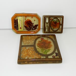 May include: Three vintage resin encased decorative items. One is an octagonal frame with a thermometer and dried flowers. Another is a square frame with an hourglass and flowers. The third is a rectangular frame with seeds and a flower.