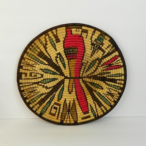 Puede incluir: Cesta redonda tejida con un diseño central rojo y patrones radiales en tonos beige, marrón, verde y rojo. La cesta tiene un borde marrón oscuro. Esta pieza decorativa muestra intrincadas técnicas de tejido.