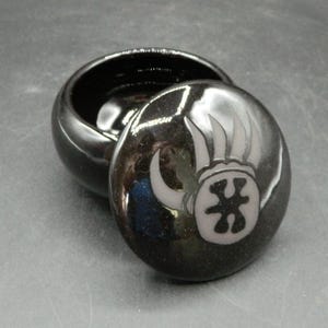 May include: A small, round, black ceramic trinket box with a lid. The lid features a gray design of a stylized emblem. The box is open, revealing the interior. The glossy finish reflects light.