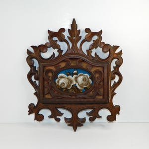 May include: A dark brown wooden wall pocket featuring an ornate, carved scrollwork frame. The centre contains an oval needlepoint panel displaying two white roses against a vibrant blue background, set within a rectangular wooden border.