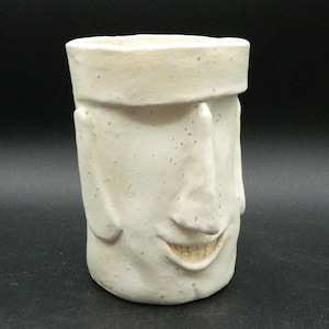 May include: A cream-colored ceramic mug shaped like a smiling face. The mug has a wide, rectangular top and a textured surface with small, dark spots. The face features a prominent nose, eyes, and a wide, toothy grin.