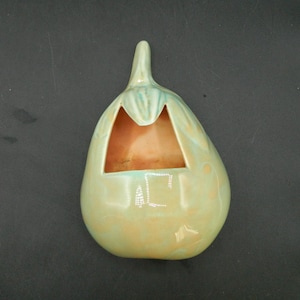 May include: A ceramic wall pocket in the shape of a pear, with a light green and peach glaze. The pear-shaped pocket has a rectangular opening and a stem. The interior is a peach color.