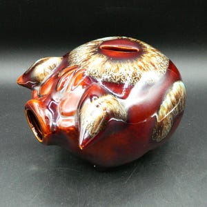 Pode incluir: Um cofrinho de cerâmica em forma de porco. O cofrinho é esmaltado em tons de marrom e bege, com uma ranhura na parte superior para inserir moedas. O porco tem orelhas pontudas e um focinho. É um item decorativo.