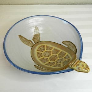 Könnte beinhalten: Eine Keramikschale mit handgemaltem Meeresschildkröten-Design. Die Schale hat einen weißen Innenraum mit blauem Rand und eine Schildkrötenillustration in Braun- und Gelbtönen. Der Kopf der Schildkröte ragt aus dem Rand heraus.