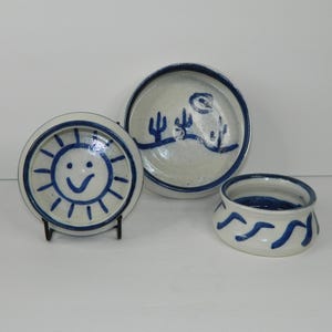 Puede incluir: Un conjunto de tres piezas de cerámica hechas a mano. Incluye un cuenco pequeño con diseño de olas azules, un plato con un sol sonriente y un plato más grande con una escena desértica, todo con detalles azules sobre fondo crema.