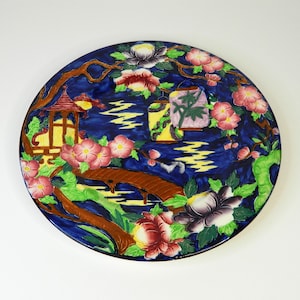 Könnte beinhalten: Ein runder Keramikteller mit dunkelblauem Hintergrund, der eine japanische Gartenszene zeigt. Das Design umfasst eine Brücke, eine Pagode, Laternen und blühende Blumen in Rosa, Rot und Weiß. Braune Äste und grünes Laub umrahmen die Szene.