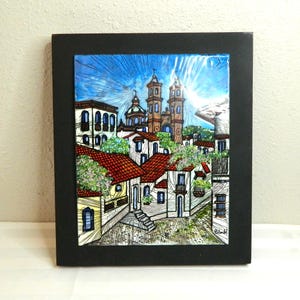 May include: A framed artwork depicting a detailed cityscape with buildings, red tile roofs, and a church under a bright blue sky. The artwork is set within a black frame, showcasing a vibrant, colorful scene.