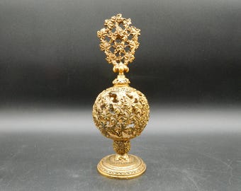 May include: Ornate gold-colored perfume bottle with a spherical body and a decorative floral design. The bottle has a detailed, openwork pattern and a matching stopper. The base is round with an intricate design. The overall height is approximately 6 inches.