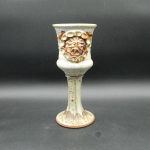 Puede incluir: Un cáliz de cerámica con un diseño de cara de sol. El cáliz es de color blanquecino con detalles en marrón y azul. La cara del sol está tallada en la parte superior. El cáliz mide aproximadamente 18 cm de altura.