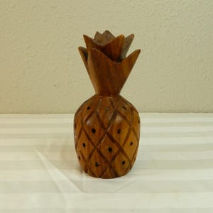 Könnte beinhalten: Eine geschnitzte Ananasfigur aus Holz. Die Ananas ist braun mit einem Rautenmuster auf dem Körper und einer stacheligen Spitze. Kleine, dunkle Punkte sind gleichmäßig in den Rautenformen verteilt. Die Figur ist ein dekorativer Gegenstand.