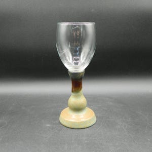 May include: A clear wine glass with a unique, handcrafted base. The base features a bulbous design with a brown and green color scheme. The glass itself is smooth and transparent, perfect for showcasing the wine's color.