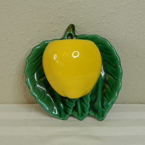 May include: A ceramic wall hanging featuring a bright yellow fruit-shaped object set on a green leaf-shaped base. The fruit has a glossy finish, and the leaf has detailed veining. The piece has a small loop for hanging.