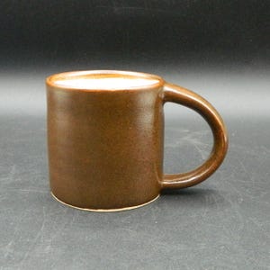 May include: A brown ceramic mug with a rounded handle. The mug has a cylindrical shape and a speckled texture. The interior of the mug is a lighter color than the exterior. The mug is suitable for hot or cold beverages.