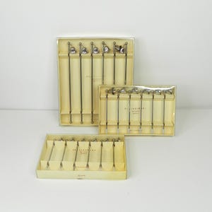 May include: Three sets of vintage cocktail picks in their original boxes. The picks have silver-toned metal stems and decorative bell-shaped tops. The boxes are a light yellow color and have the words "Party Picks" printed on them.