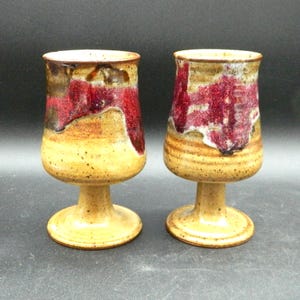 Könnte beinhalten: Zwei handgefertigte Weingläser aus Keramik. Die Gläser haben einen beigefarbenen Fuß und Stiel mit einer rot-braunen glasierten Oberseite. Die Gläser sind etwa 15 cm hoch.