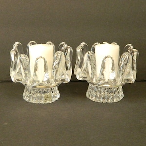 May include: Two clear glass candle holders with a unique design. Each holder features a circular base with a textured pattern and a series of curved, petal-like shapes around the top. White candles are placed in the center.