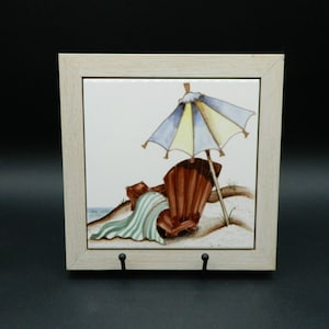 Puede incluir: Un azulejo decorativo enmarcado que representa una escena de playa con una silla Adirondack marrón, una toalla a rayas y una sombrilla azul y amarilla. El marco es de madera clara y tiene dos ganchos negros en la parte inferior.