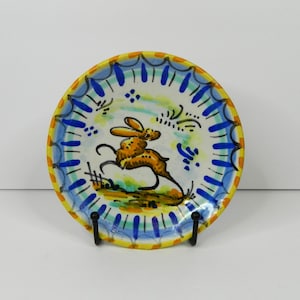 May include: A small, round decorative plate with a hand-painted design. The plate features a yellow rim, a blue and white border, and a central image of a jumping orange rabbit. The plate is displayed on a black metal stand.