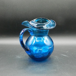 Puede incluir: Una jarra de vidrio azul cobalto con un borde rizado y un asa. La jarra está hecha de vidrio transparente con un tinte azul.