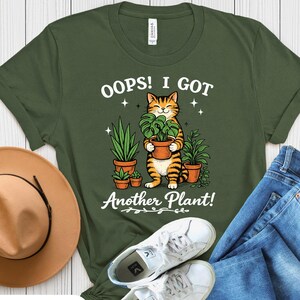May include: Military green unisex t-shirt with the text "Oops! I Got Another Plant!" featuring a cartoon cat holding a potted plant. The shirt is paired with blue jeans, white sneakers, and a brown hat.
