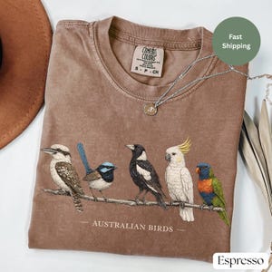 Puede incluir: Una camiseta marr&oacute;n de manga corta con un gr&aacute;fico de cinco aves australianas posadas en una rama y el texto AUSTRALIAN BIRDS impreso debajo. La prenda tiene cuello redondo y una etiqueta de la marca Comfort Colors en el interior.