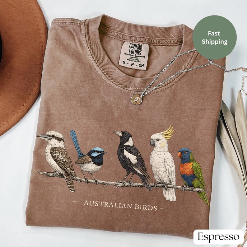 Puede incluir: Una camiseta marr&oacute;n de manga corta con un gr&aacute;fico de cinco aves australianas posadas en una rama y el texto AUSTRALIAN BIRDS impreso debajo. La prenda tiene cuello redondo y una etiqueta de la marca Comfort Colors en el interior.