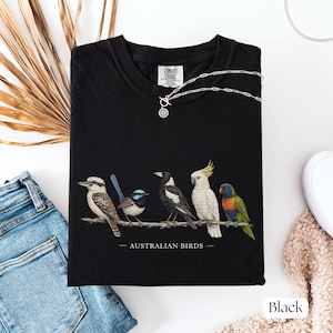 Puede incluir: Una camiseta negra con un gr&aacute;fico detallado de cinco aves australianas posadas en una rama, incluyendo un kookaburra, un maluro, una urraca, una cacat&uacute;a y un lori, con el texto AUSTRALIAN BIRDS impreso debajo de la ilustraci&oacute;n.