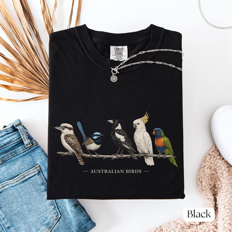 Puede incluir: Una camiseta negra con un gr&aacute;fico detallado de cinco aves australianas posadas en una rama, incluyendo un kookaburra, un maluro, una urraca, una cacat&uacute;a y un lori, con el texto AUSTRALIAN BIRDS impreso debajo de la ilustraci&oacute;n.