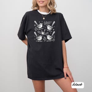 May include: Black t-shirt featuring a detailed white illustration of several birds perched on branches with leaves. The design includes small stars and is printed on a black, oversized tee. The word "black" is in the bottom right corner.