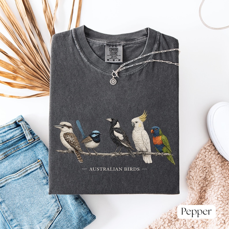 Puede incluir: Una camiseta de manga corta gris carb&oacute;n con un estampado gr&aacute;fico de cinco aves australianas posadas en una rama y el texto "AUSTRALIAN BIRDS" impreso debajo. La camiseta tiene cuello redondo y una etiqueta de la marca Comfort Colors.