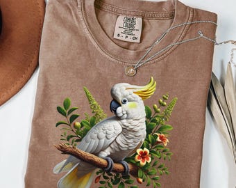 Może przedstawiać: Brązowa koszulka Comfort Colors z szczegółową ilustracją białej kakadu siedzącej na gałęzi, otoczonej zielonymi liśćmi i czerwonymi kwiatami. Koszulka ma okrągły dekolt i krótkie rękawy. Srebrny naszyjnik z wisiorkiem jest zarzucony na koszulkę.