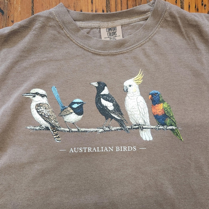 Puede incluir: Una camiseta color topo con un gr&aacute;fico de cinco aves australianas posadas en una rama. Las aves incluyen un cucaburra, un maluro, una urraca, una cacat&uacute;a y un lori. El texto "AUSTRALIAN BIRDS" est&aacute; impreso debajo de la rama.