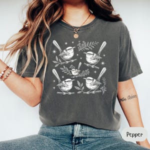 May include: A dark gray t-shirt featuring a detailed white illustration of several birds perched on branches with leaves. The birds are surrounded by small star-like accents. The shirt is a comfortable, casual style.
