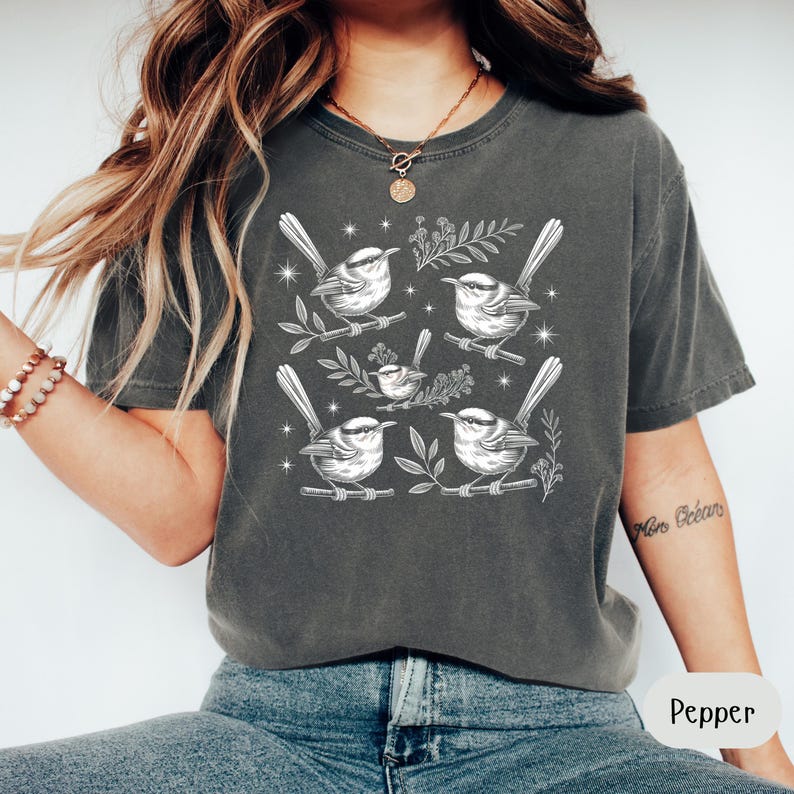 May include: A dark gray t-shirt featuring a detailed white illustration of several birds perched on branches with leaves. The birds are surrounded by small star-like accents. The shirt is a comfortable, casual style.