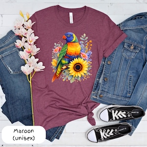 May include: Maroon unisex t-shirt with a vibrant parrot design, set amongst sunflowers and floral elements. The parrot's plumage includes blue, green, yellow, and orange. The shirt is paired with denim jeans, a denim jacket, and black trainers.