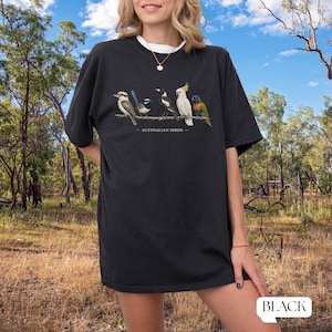 Puede incluir: Una camiseta negra de manga corta con una ilustraci&oacute;n gr&aacute;fica de cinco aves australianas posadas en una rama con el texto AUSTRALIAN BIRDS impreso debajo. Las aves incluyen un cucaburra, una ratona australiana, una urraca, una cacat&uacute;a y un lori.