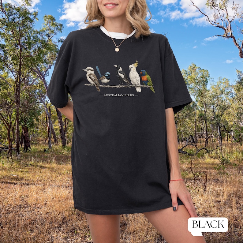 Puede incluir: Una camiseta negra de manga corta con una ilustraci&oacute;n gr&aacute;fica de cinco aves australianas posadas en una rama con el texto AUSTRALIAN BIRDS impreso debajo. Las aves incluyen un cucaburra, una ratona australiana, una urraca, una cacat&uacute;a y un lori.