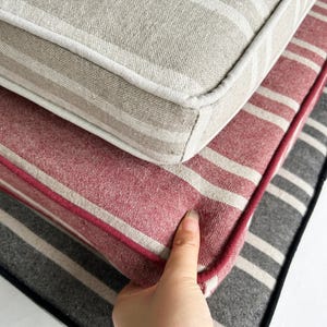 May include: Close-up of stacked striped cushions in neutral tones. The top cushion is beige with white stripes, the middle one is red with white stripes, and the bottom one is gray with white stripes. A hand is touching the red cushion.