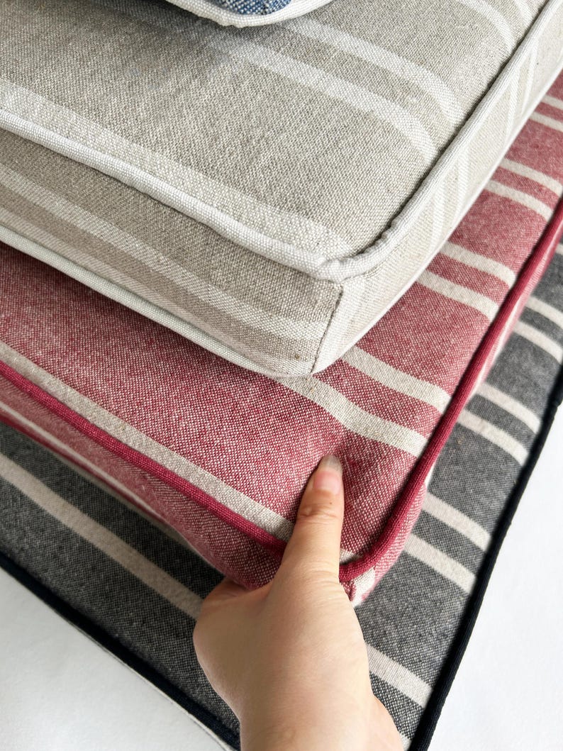 May include: Close-up of stacked striped cushions in neutral tones. The top cushion is beige with white stripes, the middle one is red with white stripes, and the bottom one is gray with white stripes. A hand is touching the red cushion.