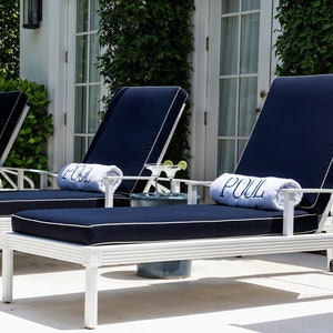 Könnte beinhalten: Zwei weiße Liegestühle mit blauen Kissen und weißen Handtüchern, auf denen "POOL" steht. Die Stühle stehen auf einer weißen Terrasse mit einem kleinen Tisch und einem Glas Limettensaft.