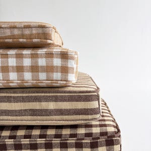 Op de afbeelding: Een stapel van vier vierkante kussens in verschillende patronen en bruintinten. Het bovenste kussen is effen bruin, gevolgd door een wit en bruin ruitpatroon, dan een bruin en beige streepjespatroon, en ten slotte een groot bruin en beige ruitpatroon.