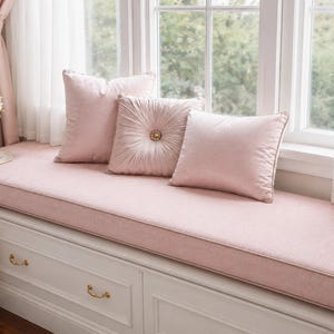 May include: A window seat featuring a long, light pink cushion atop a white storage bench with gold handles. Three decorative pillows in matching light pink shades are arranged on the cushion, including one square pillow with a central button detail.