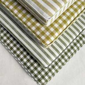 May include: Four square cushions in shades of green and white. The top cushion has a striped pattern, the second a gingham check, the third a gingham check, and the bottom a larger gingham check. The cushions are stacked at an angle.
